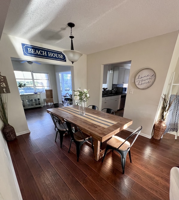 A dining area with a wooden table, chairs, and a vase of flowers. There is a sign that reads "BEACH HOUSE".