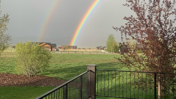 A wooden deck with a table and chairs, overlooking a grassy field with houses in the distance and a rainbow in the sky.