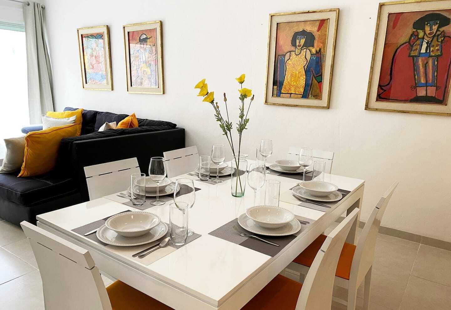Un coin repas avec une table blanche dressée pour quatre personnes, entourée de chaises, et des œuvres d’art accrochées aux murs.