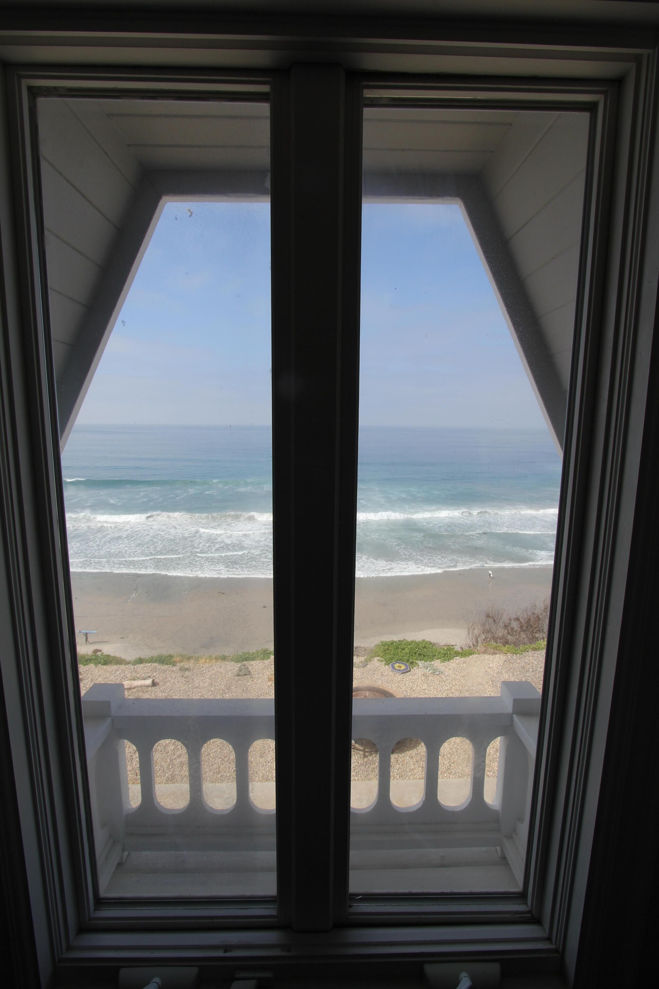 Uitzicht vanuit een raam met balkon op een strand met golven en een heldere lucht.