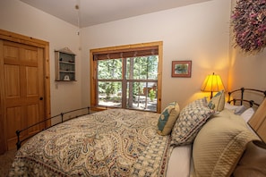 Ein Schlafzimmer mit einem großen Bett, einer Holztür, einem Fenster mit Blick auf Bäume, einer Nachttischlampe und einem Regal mit Dekoration.