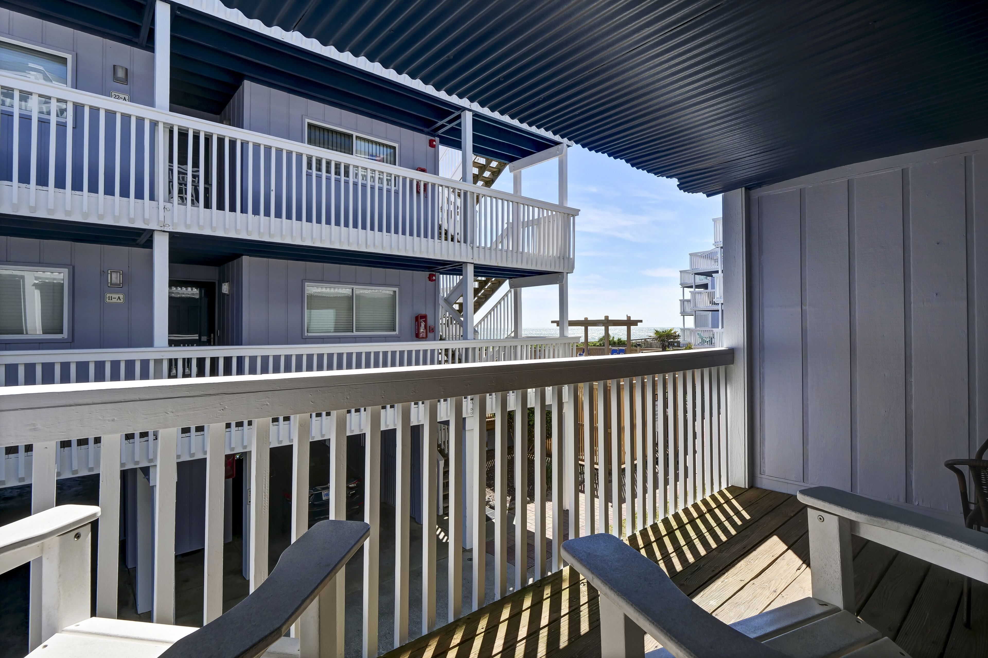 Un balcon à plusieurs niveaux avec des balustrades blanches, un espace couvert doté d’un toit en tôle ondulée et une terrasse en bois avec des bancs.
