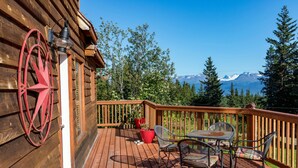 A wooden deck with a table and chairs, a red metal decoration, and a view of mountains. - Moose Meadows near Kilcher Homestead in Alaska the Last Frontier (Homer)