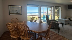 A dining area with a wooden table and chairs, large windows with a view of the ocean, and a living area with a sofa and a TV. - Starr Creek - Ocean and Creek right outside! (Yachats)