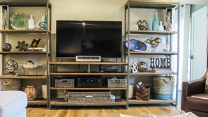 A living room with a large flat-screen TV on a wooden stand, surrounded by shelves with various decorative items and storage boxes. - Immerse Yourself in Wine Country Long Term Okay (Yountville)