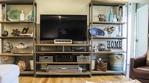 Uma sala de estar com uma televisão de tela plana grande em um suporte de madeira, cercada por estantes com diversos objetos decorativos e caixas organizadoras.
