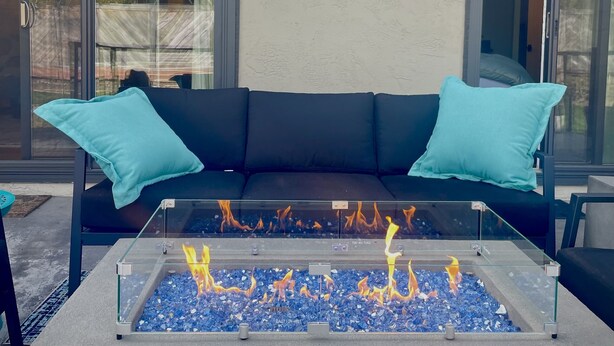 A glass-top fire pit with blue stones, a black outdoor sofa with teal cushions, and a glass door leading to a patio area. - Immerse Yourself in Wine Country Long Term Okay (Yountville)