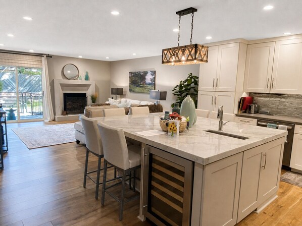 A modern kitchen with a central island, bar stools, and a dining area with a large window.