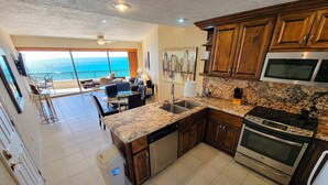 A kitchen with granite countertops, stainless steel appliances, and wooden cabinets. There is a dining area with a view of the ocean.