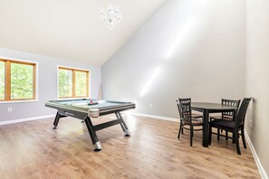 A room with a pool table, a dining table with chairs, and large windows with a view of trees.