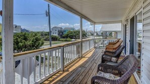 A covered porch with wicker chairs, a wooden deck, and a view of houses and greenery. - Sassy Seahorse | Pet Friendly + Near Area Fun! (Surf City)