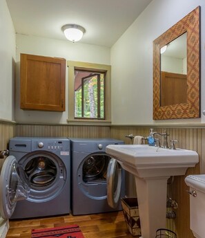 A laundry room with two front-loading washing machines, a sink, and a window with a view of trees. - Tree House Feel in a Perfect Location<br> (Lake Placid)