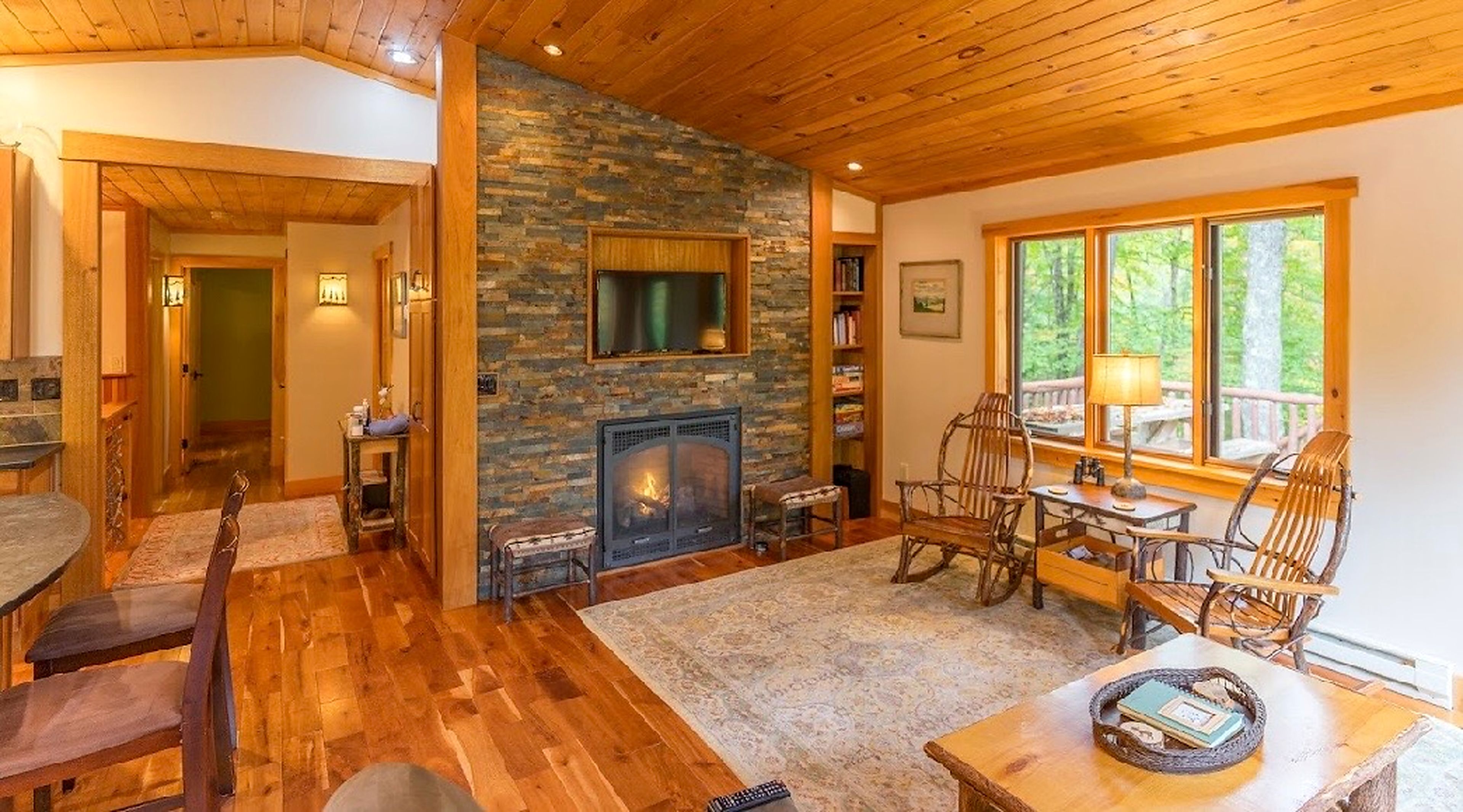 A cozy living room with a stone fireplace, wooden flooring, and a large window offering a view of the outdoors.