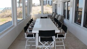 Ein überdachter Essbereich im Freien mit einem langen Tisch, Stühlen, großen Fenstern und Blick auf den Strand.