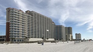 Een groot hotel met meerdere verdiepingen en balkons, gelegen aan een zandstrand onder een heldere lucht.