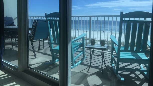 Een balkon met turquoise stoelen en een kleine tafel, met uitzicht op een strand waar mensen in de verte lopen.