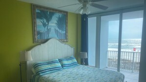 Een slaapkamer met een bed, een plafondventilator, een schilderij en uitzicht op het strand door het raam.