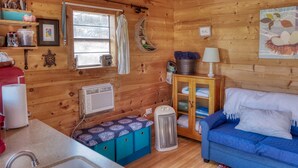 A cozy cabin interior with a kitchenette, a blue sofa, a wooden cabinet, and a window with curtains.