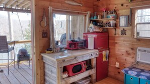 A compact kitchen with wooden walls, a built-in microwave, a red refrigerator, and a window overlooking a forested area.