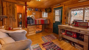A cozy cabin interior with a wooden interior, a bed, a kitchen area with a microwave, and a window with a view of trees.