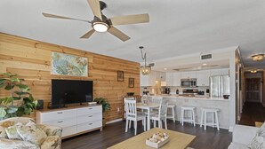 Una sala de estar moderna con televisor, paneles de madera en la pared, ventilador de techo y un comedor con sillas blancas, con una cocina al fondo.