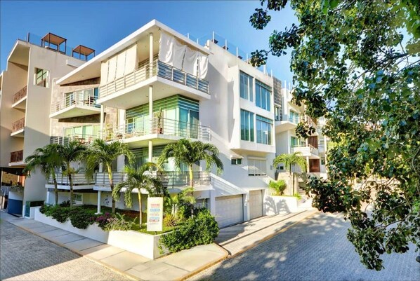 A multi-story hotel with balconies, a covered outdoor area, and a garden with palm trees.