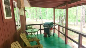 A covered outdoor area with a green deck, two Adirondack chairs, a barbecue grill, and a table. - Cabin # 2, Kentucky Lake, Bass Fishing, Camden, Tn, (Nashville 1, Memphis 1.5) (Camden)