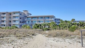 A beachfront hotel with multiple balconies, a sandy path leading to the entrance, and a clear blue sky.
