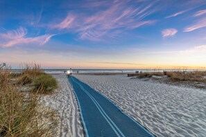 Un sentier bordant la plage, avec un tapis bleu qui mène à l’eau, entouré de sable et d’herbe.