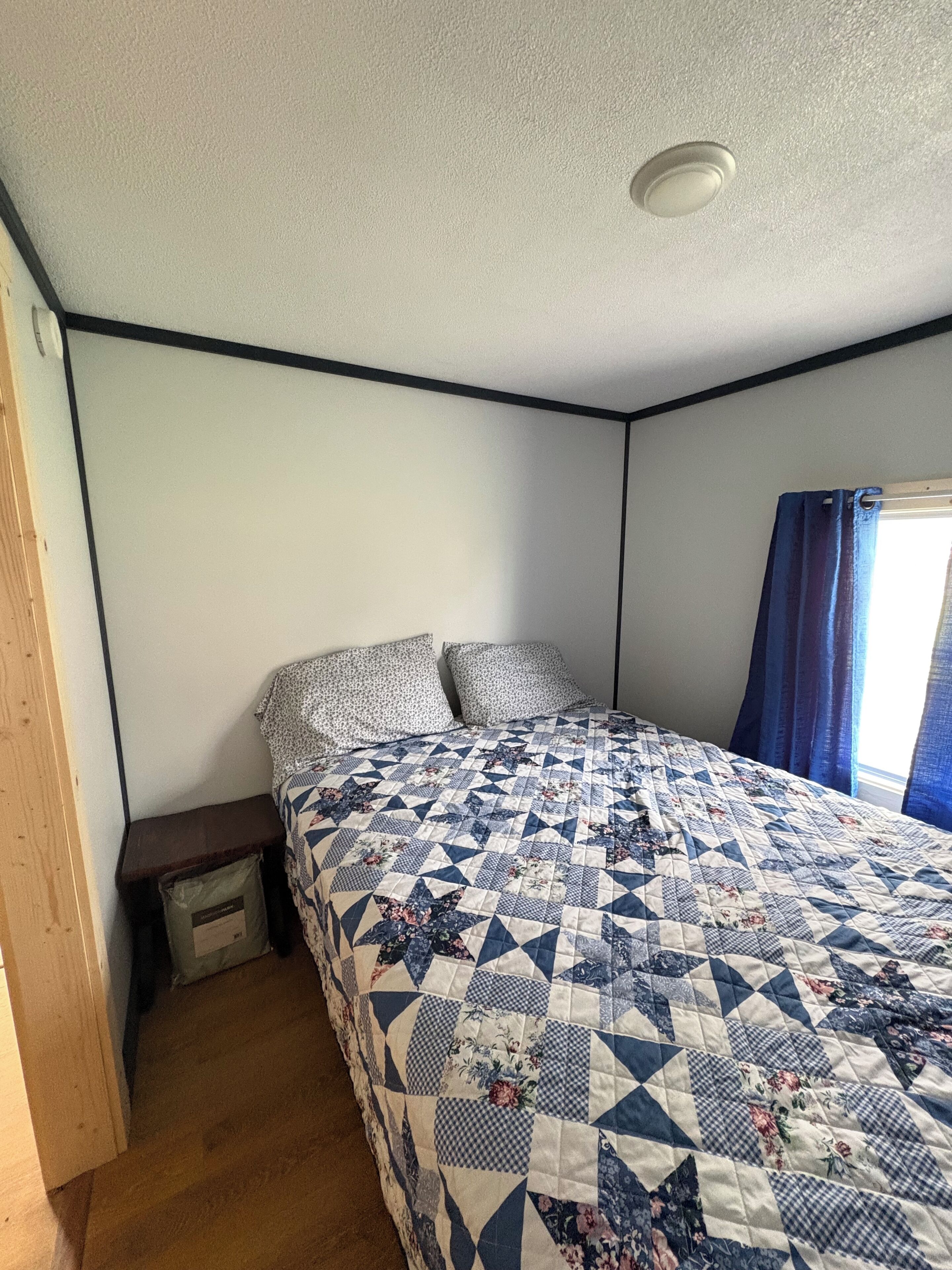 Un dormitorio con cama, mesita de noche, piso de madera, luz en el techo y ventana con cortinas.