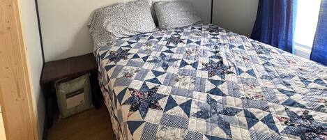 Un dormitorio con cama, mesita de noche, piso de madera, luz en el techo y ventana con cortinas.