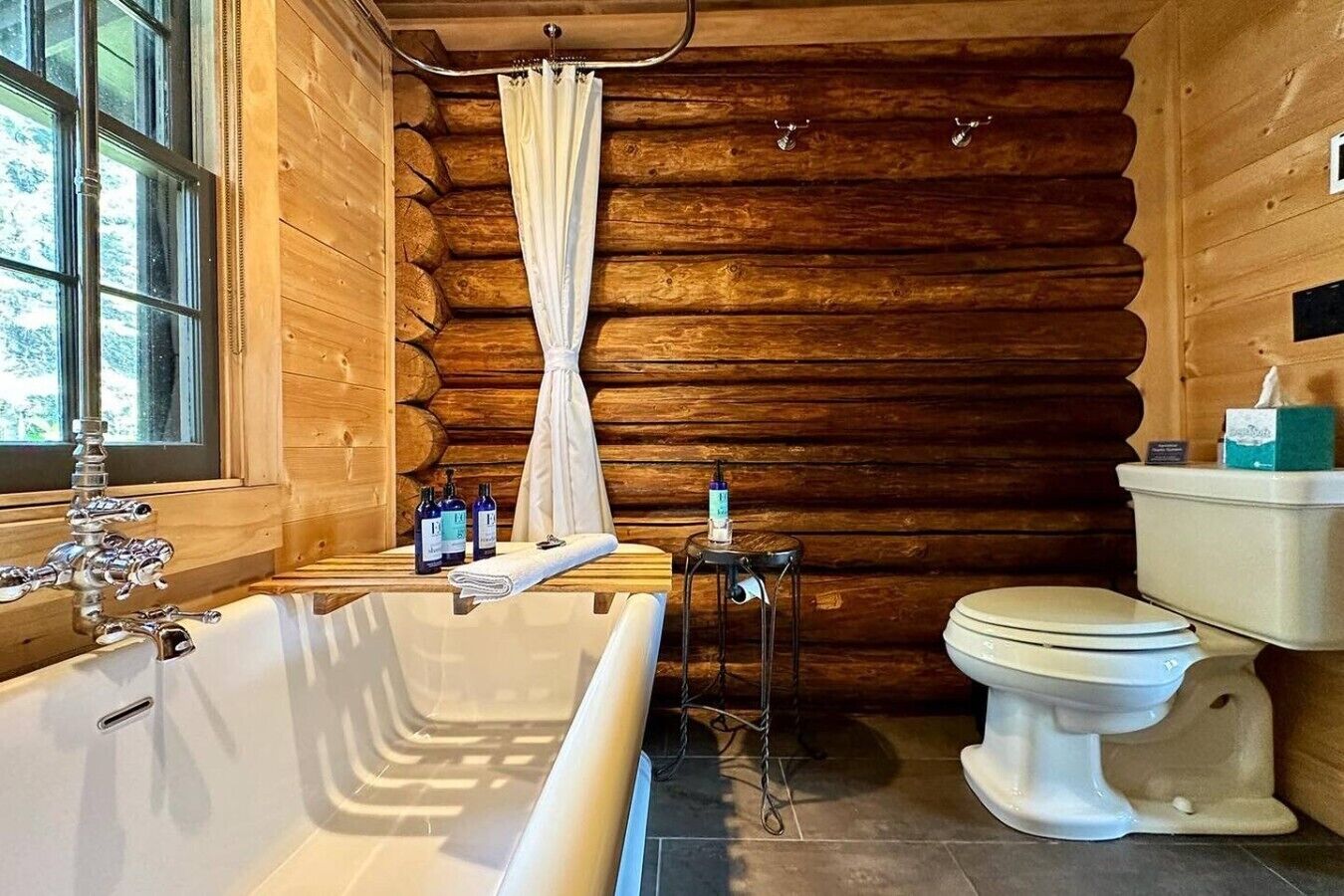 A log cabin bathroom with a bathtub, toilet, and window.