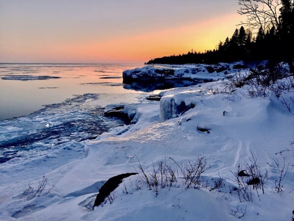 A snowy landscape with a frozen body of water, ice formations, and a forested area.
