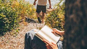Una persona camina por un sendero junto al mar, otra persona lee un libro y hay un árbol en primer plano.