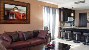 Un salon avec un canapé en cuir marron, une table basse agrémentée d’objets décoratifs, un coin cuisine avec des meubles sombres et un micro-ondes blanc, et un ventilateur de plafond.