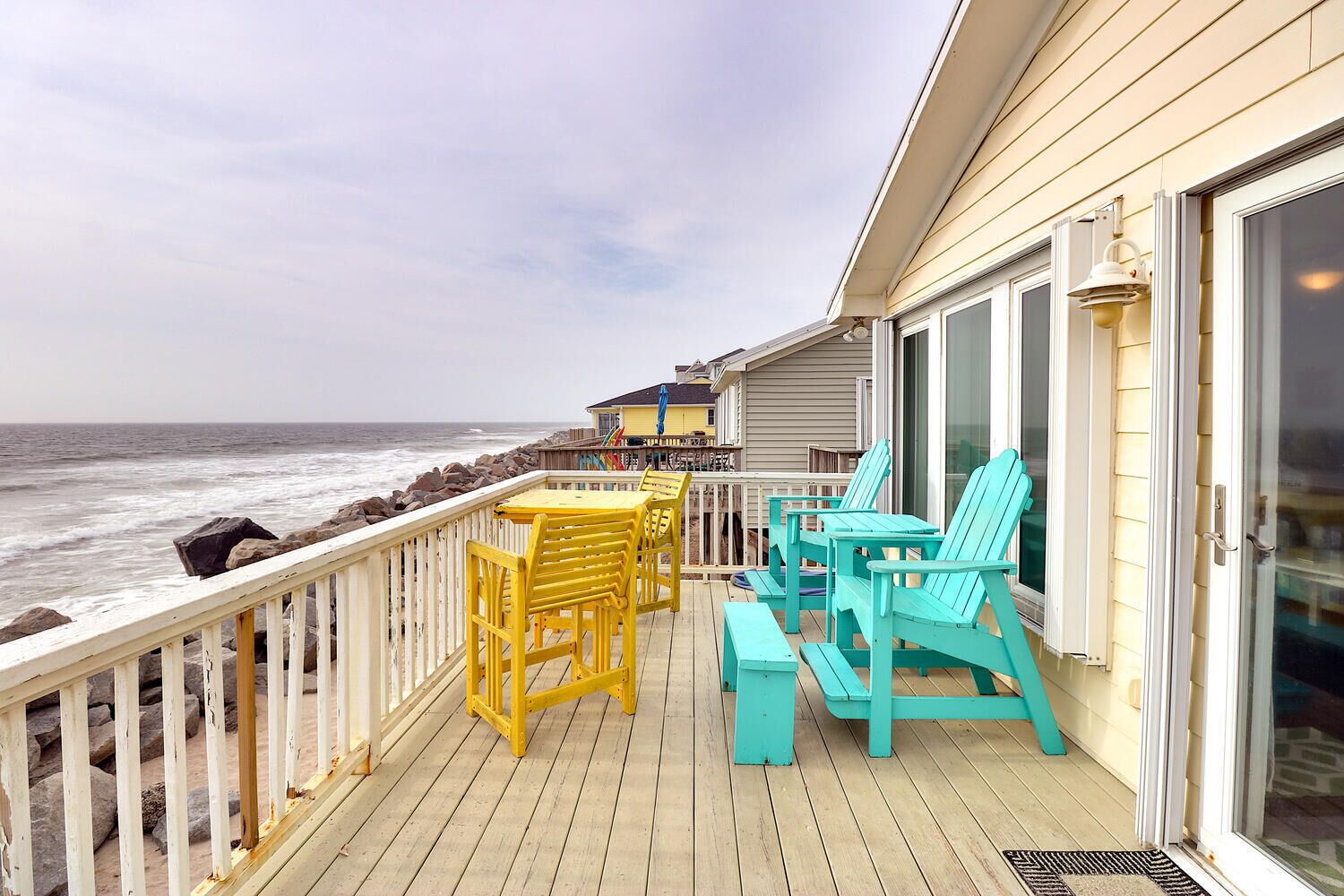 A wooden deck with yellow and turquoise chairs, a small table, and a view of the ocean.