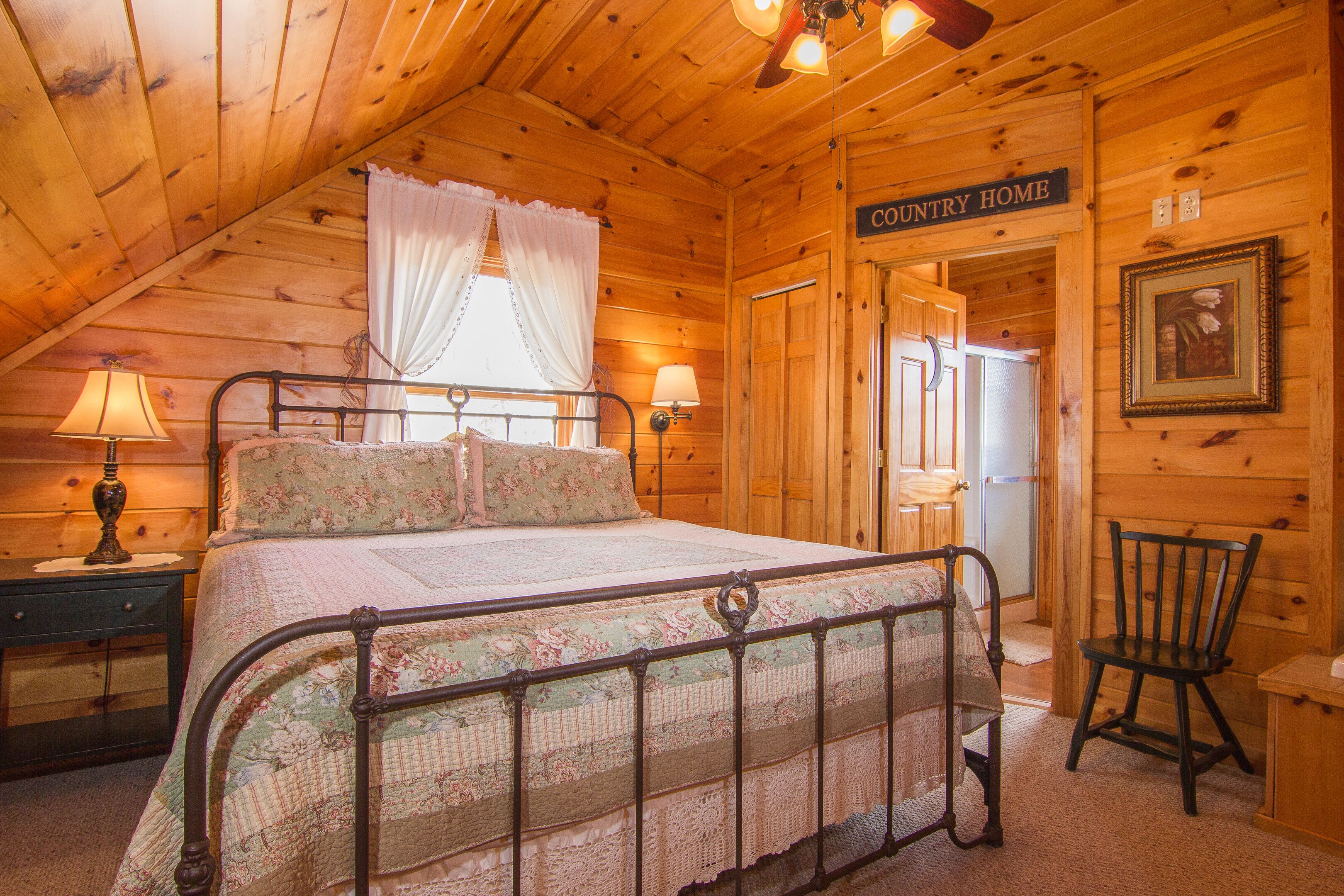 Ein gemütliches Schlafzimmer mit Holzbalkendecke, einem Metallbettgestell, einem Nachttischchen mit Lampe und einem Sessel. Ein Schild an der Wand zeigt "COUNTRY HOME".