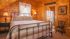 Un dormitorio acogedor con techo de madera, estructura de cama de metal, mesita de noche con lámpara y una silla. Un cartel en la pared dice “COUNTRY HOME”.