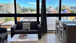 Een moderne woonkamer met een bank, salontafel en een eethoek, met grote ramen en uitzicht op de stad.