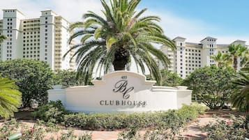 A Clubhouse sign with a palm tree in front, surrounded by greenery and a pathway.
