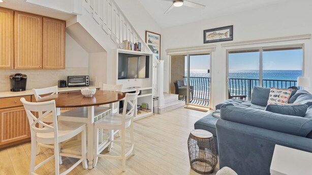 A spacious living area with a balcony overlooking the sea, a kitchen with white cabinetry, and a cozy seating area with a blue sofa.