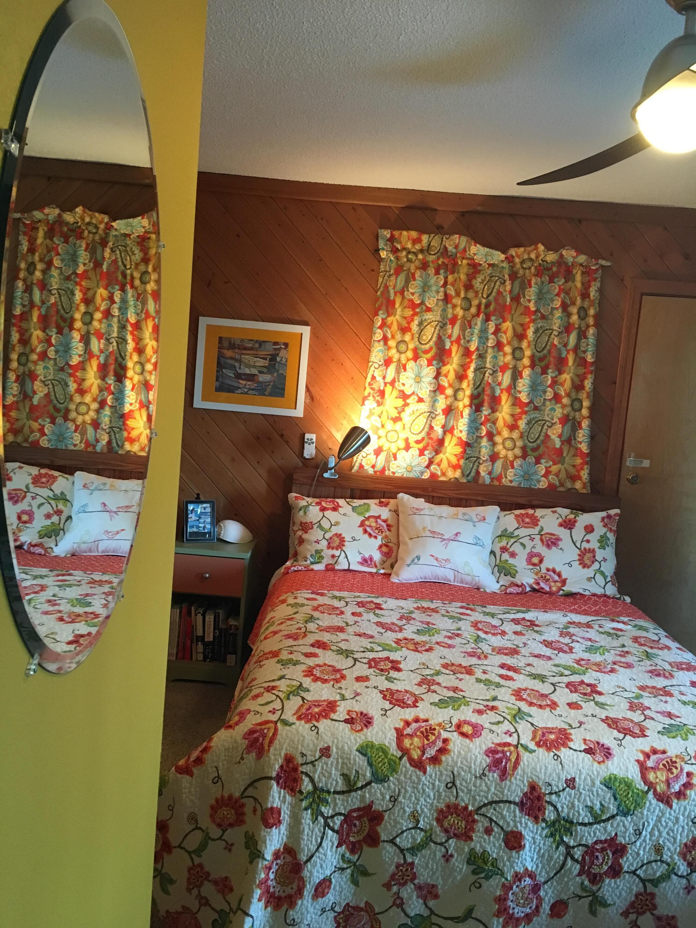 Een slaapkamer met een houten hoofdbord, bloemen gordijnen, een bed met bijpassende kussens, een nachtkastje met een lamp en een lijst met een afbeelding aan de muur.