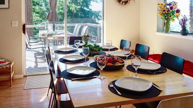 A dining area with a long table set for six, a bowl of fruit, and a vase of flowers on the windowsill. - Fir Mountain Escape - Stunning Views, River & Mountain Views, Hot Tub (Hood River)