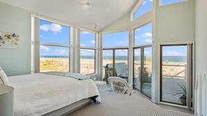 A bedroom with a large bed, a chair, and a view of the beach through large windows. - Stunning Oceanfront Home on Quiet Lane with Bay and Ocean Views. Hot tub! (Beach Haven)