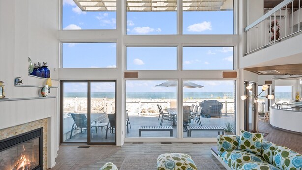 A spacious living room with a fireplace, large windows overlooking the beach, and a modern kitchen area. - Stunning Oceanfront Home on Quiet Lane with Bay and Ocean Views. Hot tub! (Beach Haven)