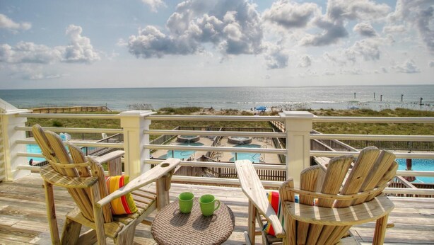 A wooden deck with wicker chairs and a small round table, overlooking a beach and a pool area.