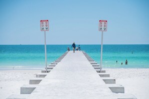A pier leading to a beach with clear blue water, people walking, and two attention signs.