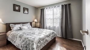 Ein Schlafzimmer mit einem großen Bett, zwei Nachttischen mit Lampen, einem Fenster mit Vorhängen und gerahmten Bildern an der Wand.