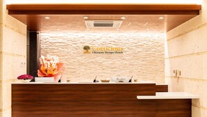 A reception desk with a wooden finish, a stone wall backdrop, and a sign for Okinawa Monpa Hotels. - Cococious Monpa (Chatan)