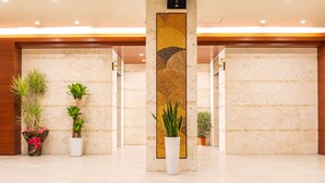 A spacious lobby with a polished marble floor, a tall pillar with a decorative gold and black artwork, and several potted plants. - Cococious Monpa (Chatan)